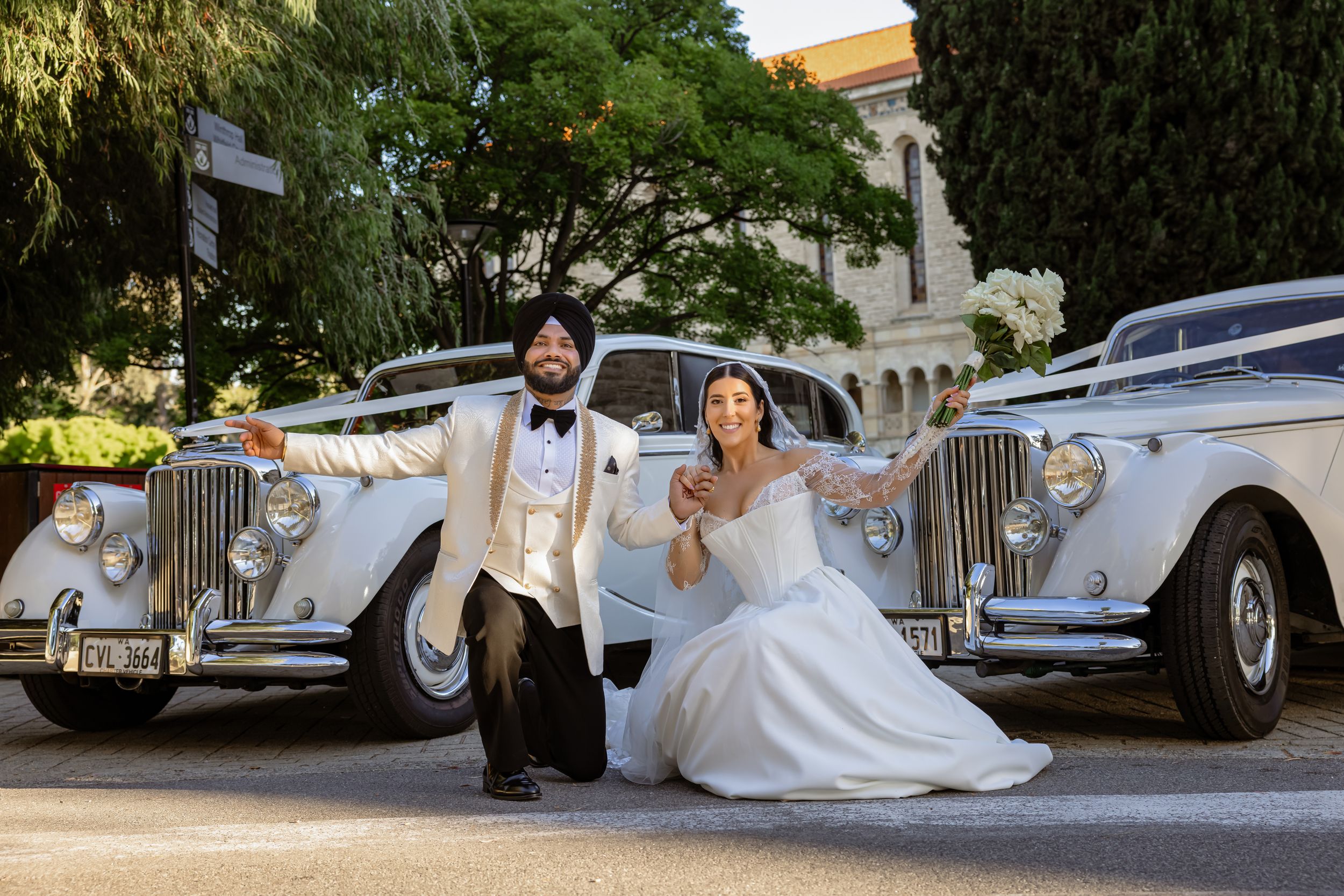 Sikh groom and bride in white wedding dress pose joyfully between two classic vintage white Rolls-Royce cars outdoors.