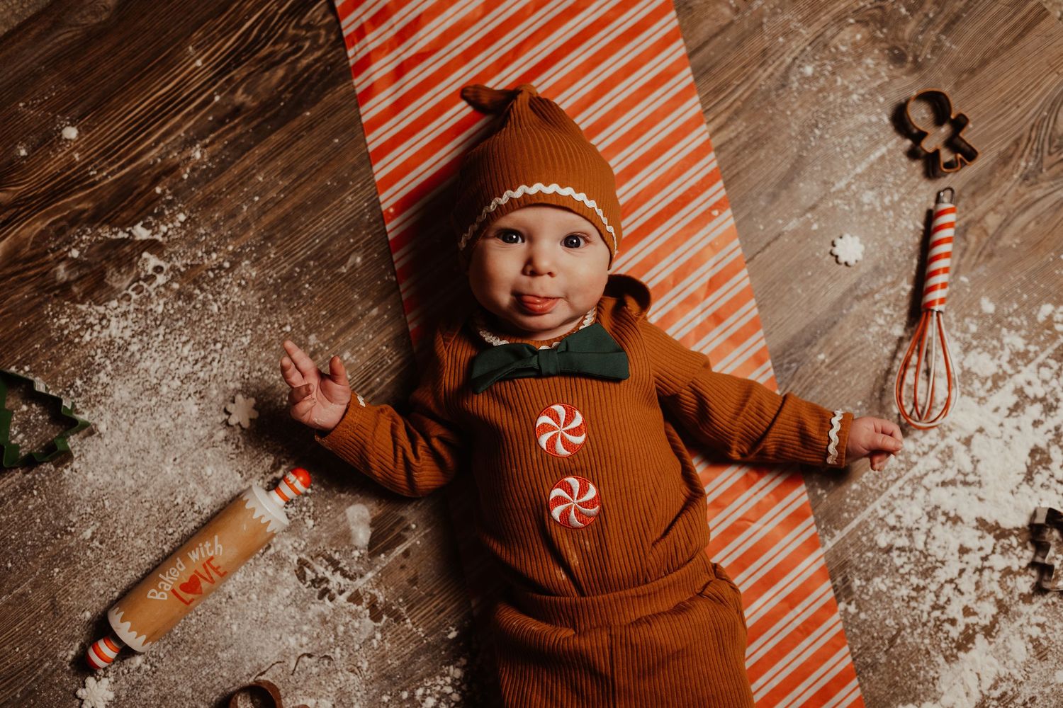 Baby dressed as gingerbread man costume lying on flour-dusted floor with candy cane, rolling pin, and whisk.