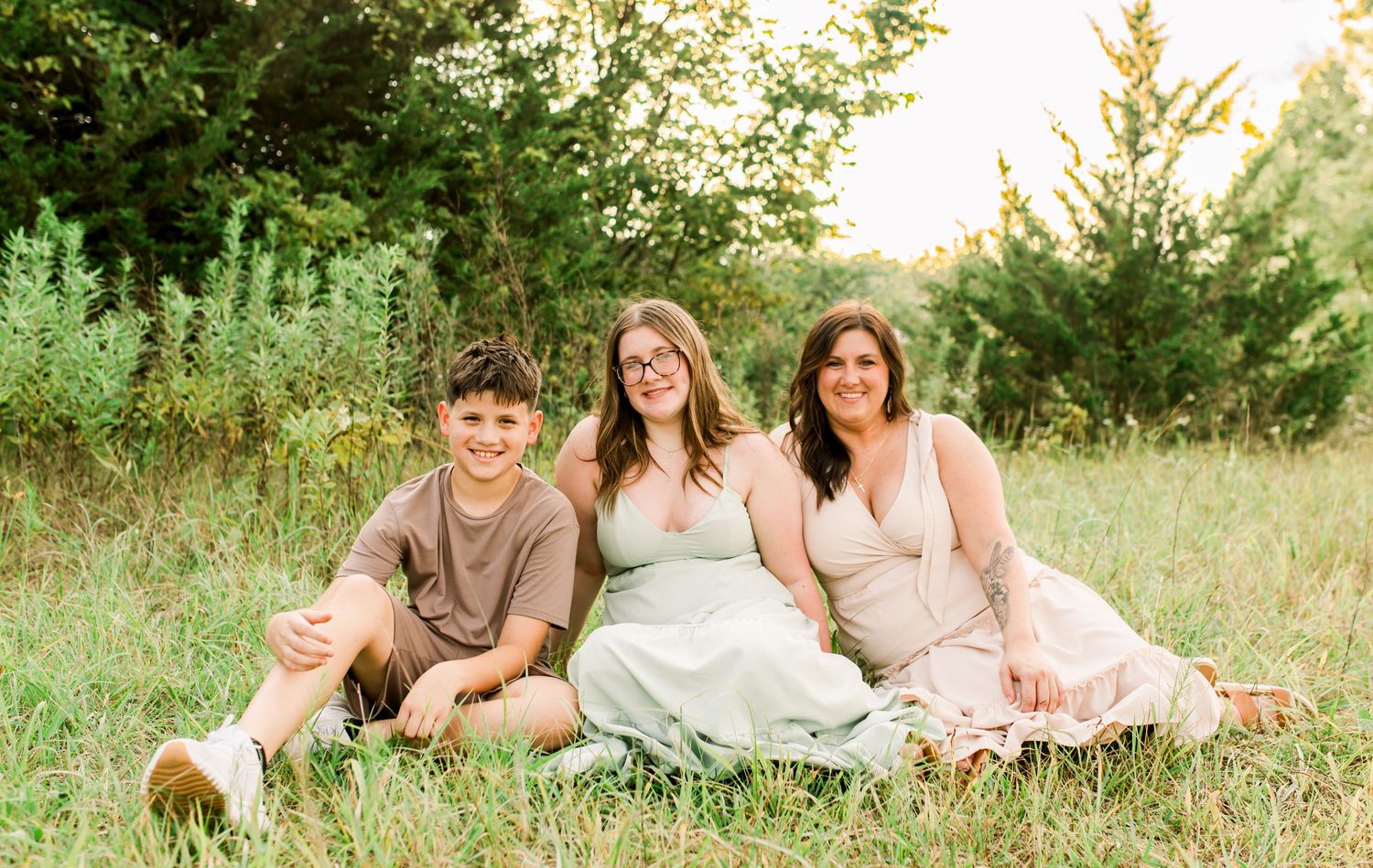 A boy and teenage girl smile at each other in a sunlit meadow, wearing brown and mint green outfits.