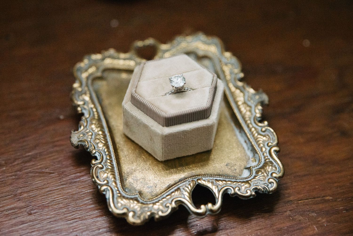 An engagement ring sits in a velvet hexagonal box on an ornate silver tray.