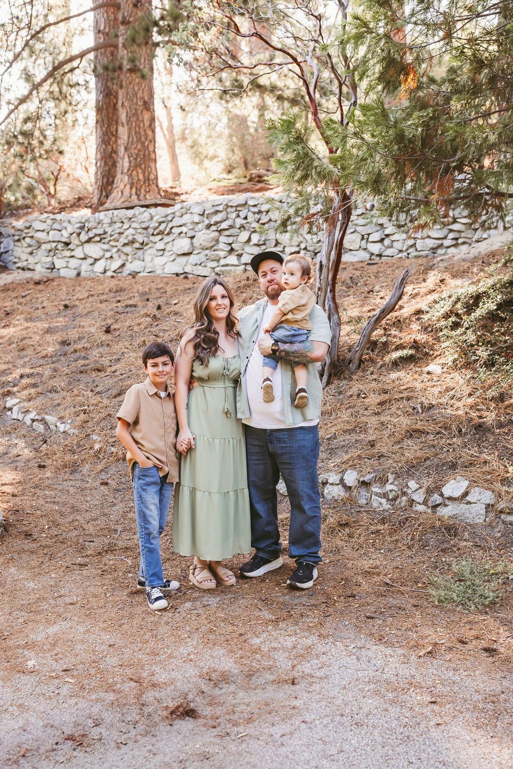 Family of four posing outdoors among pine trees and stone wall, wearing coordinated earth-tone outfits in the inland empire mountains