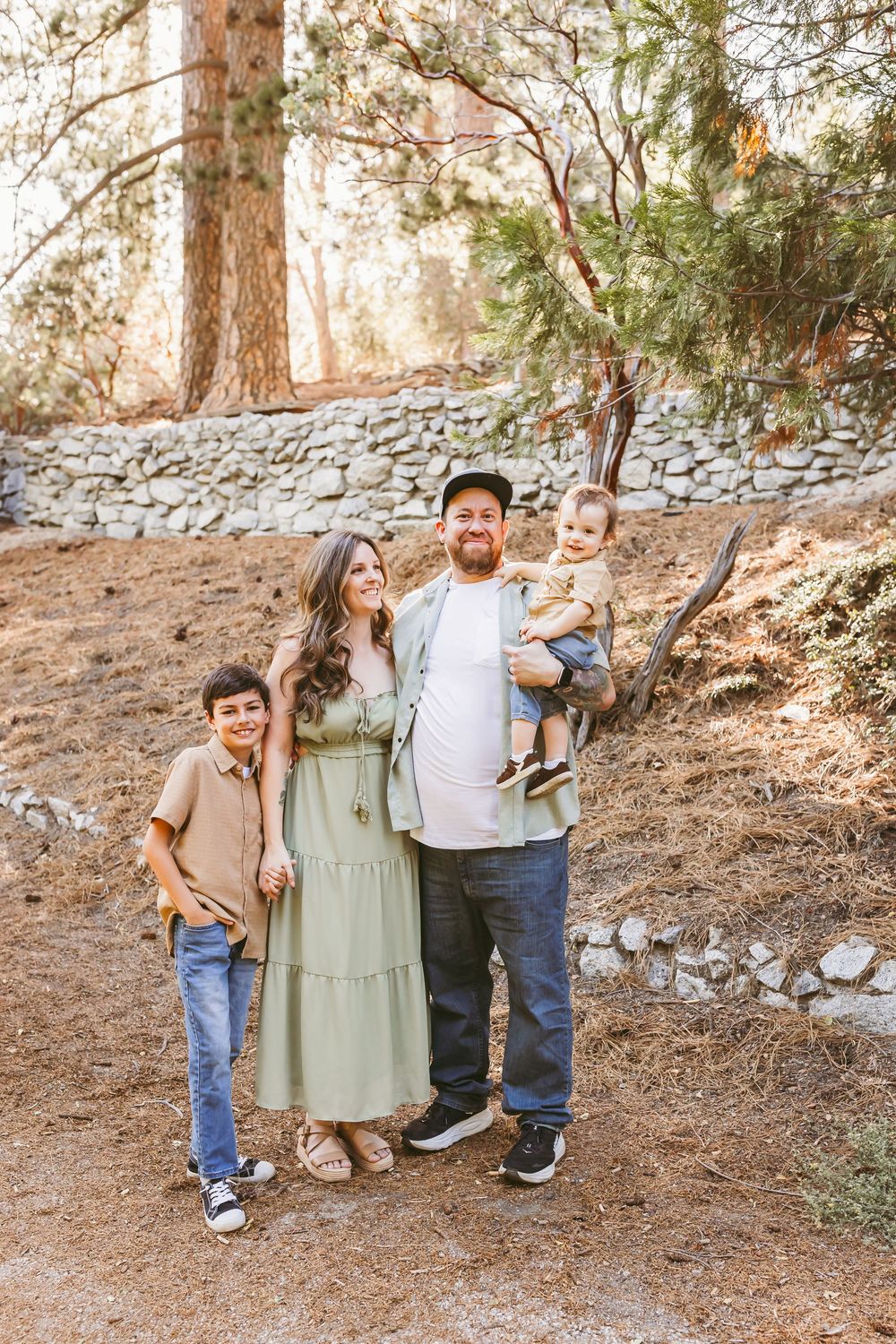 Happy family of four posing outdoors in a forest setting, parents with two young children among tall trees.