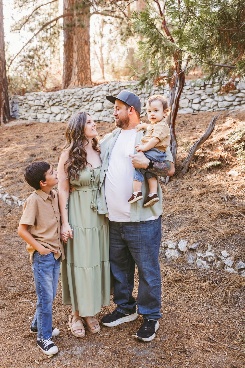 Family of four posing outdoors in a forest setting, parents and two young boys wearing casual earth-tone outfits.