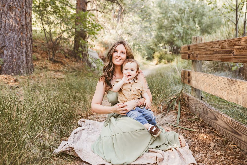 A mother in a green dress sits on a blanket outdoors holding her baby boy near a wooden fence.