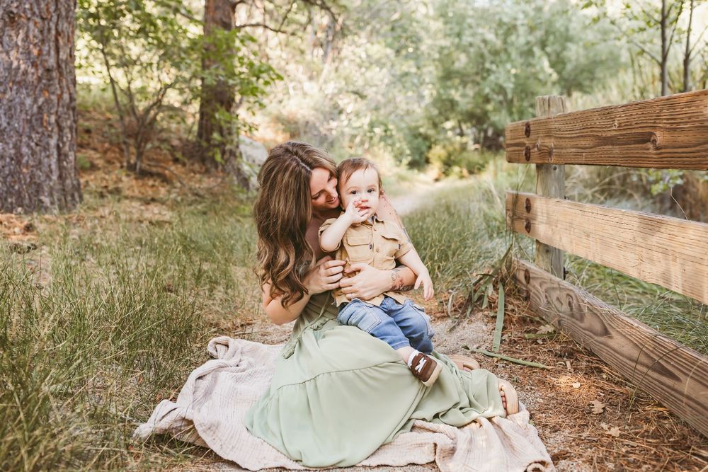 A mother in a green dress sits on a blanket outdoors, lovingly holding her baby near a wooden fence.