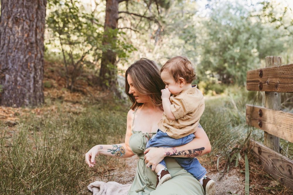 A mother holds her toddler outdoors in a lush forest setting, both smiling warmly in natural light.