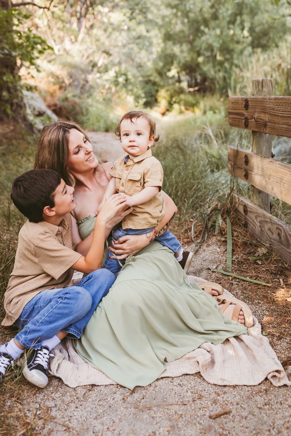 A mother sits on a blanket outdoors with her two young sons near a wooden fence surrounded by greenery.