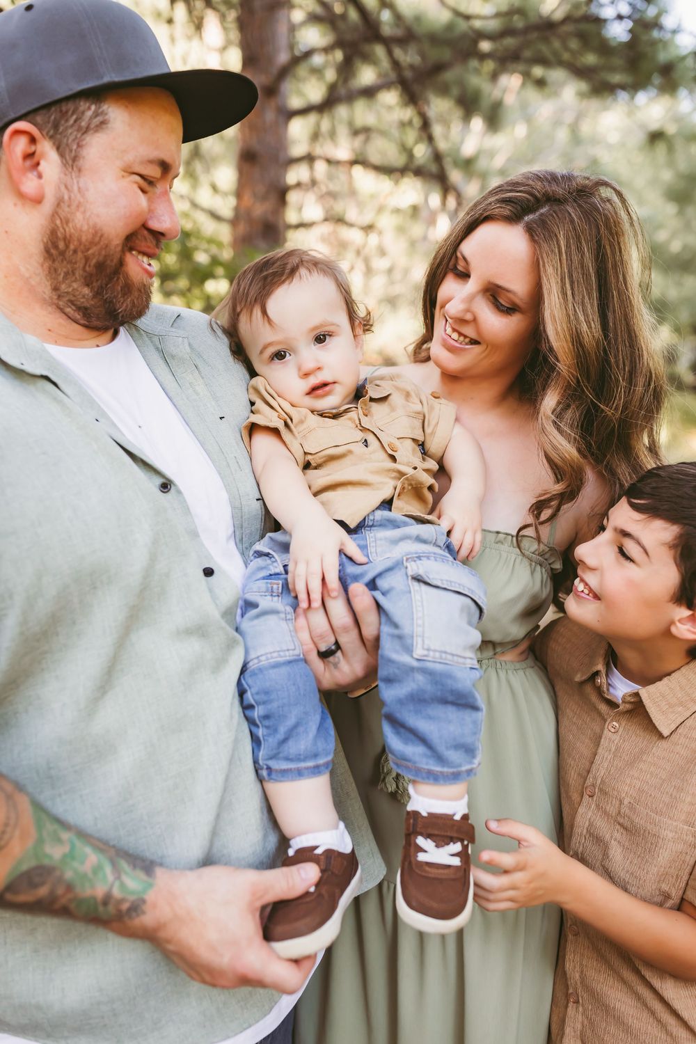 Happy family of four posing outdoors in a forest setting, parents holding young toddler while older boy smiles nearby.