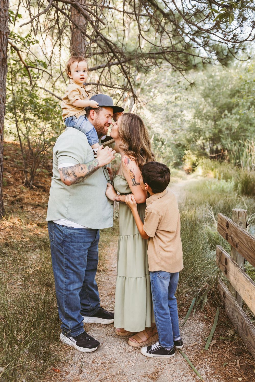 Family of four posing outdoors on a nature path, parents sharing a tender moment while holding their two young children.