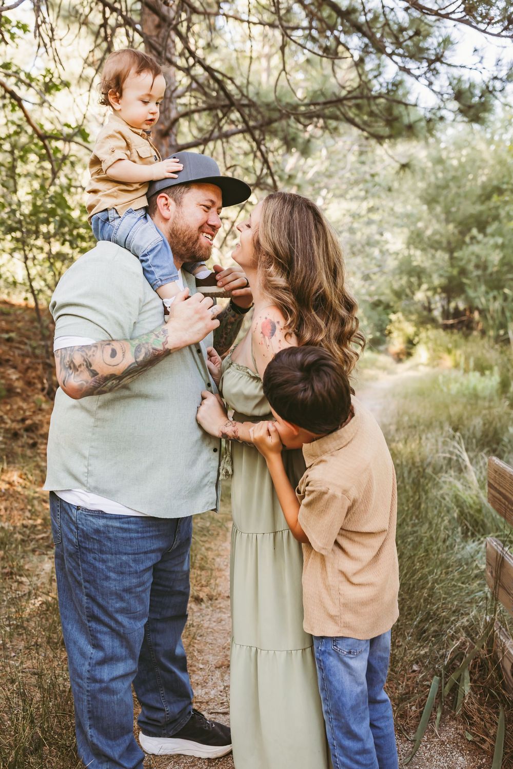 Family of four enjoying a nature walk, dad with tattoos lifts toddler on shoulders while mom and older son embrace outdoors.