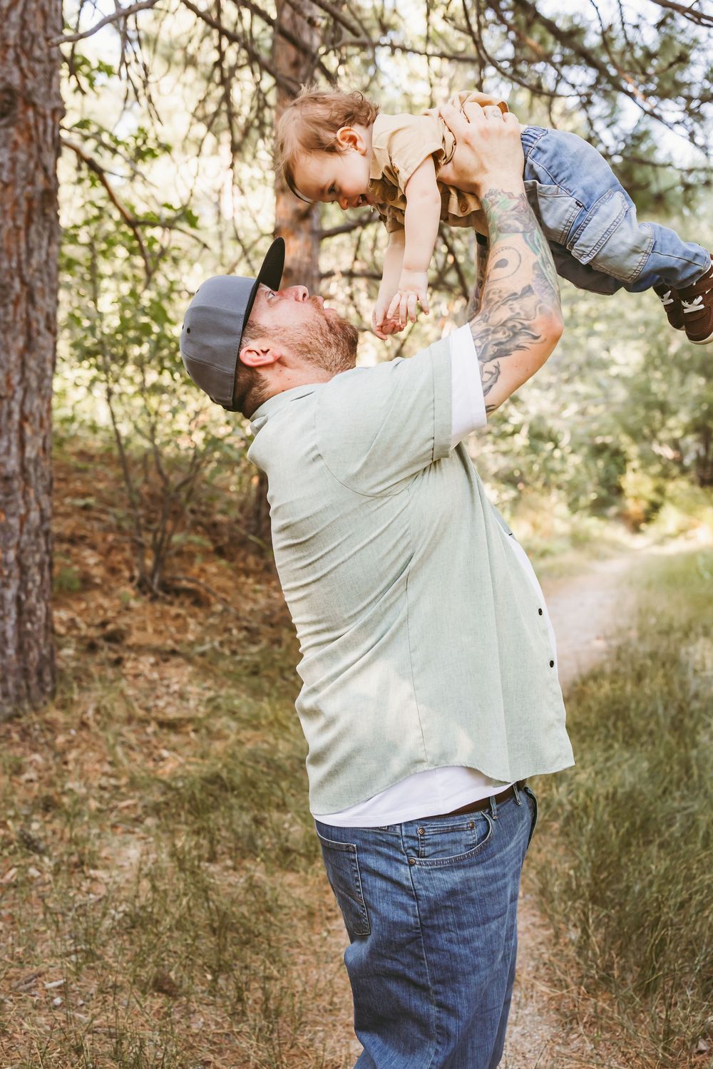 Tattooed dad joyfully lifts smiling toddler overhead on a scenic forest trail on a sunny day.