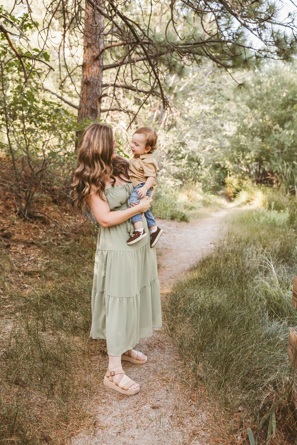 A woman in a sage green dress holds a toddler on a forest path surrounded by lush greenery.