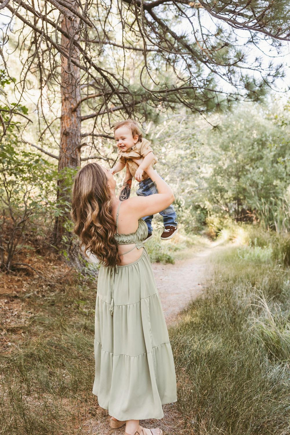 A mother in a sage green dress lifts her laughing baby outdoors on a forest path surrounded by lush greenery.