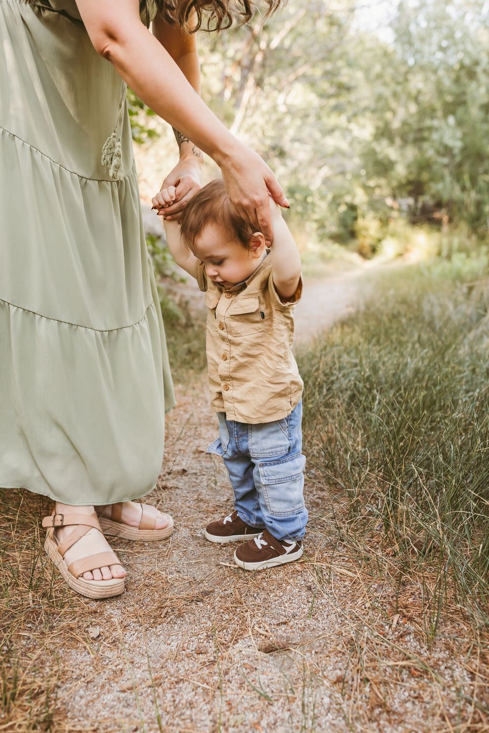 A mother in a sage green dress helps her toddler take first steps on a nature path, both wearing sandals outdoors.