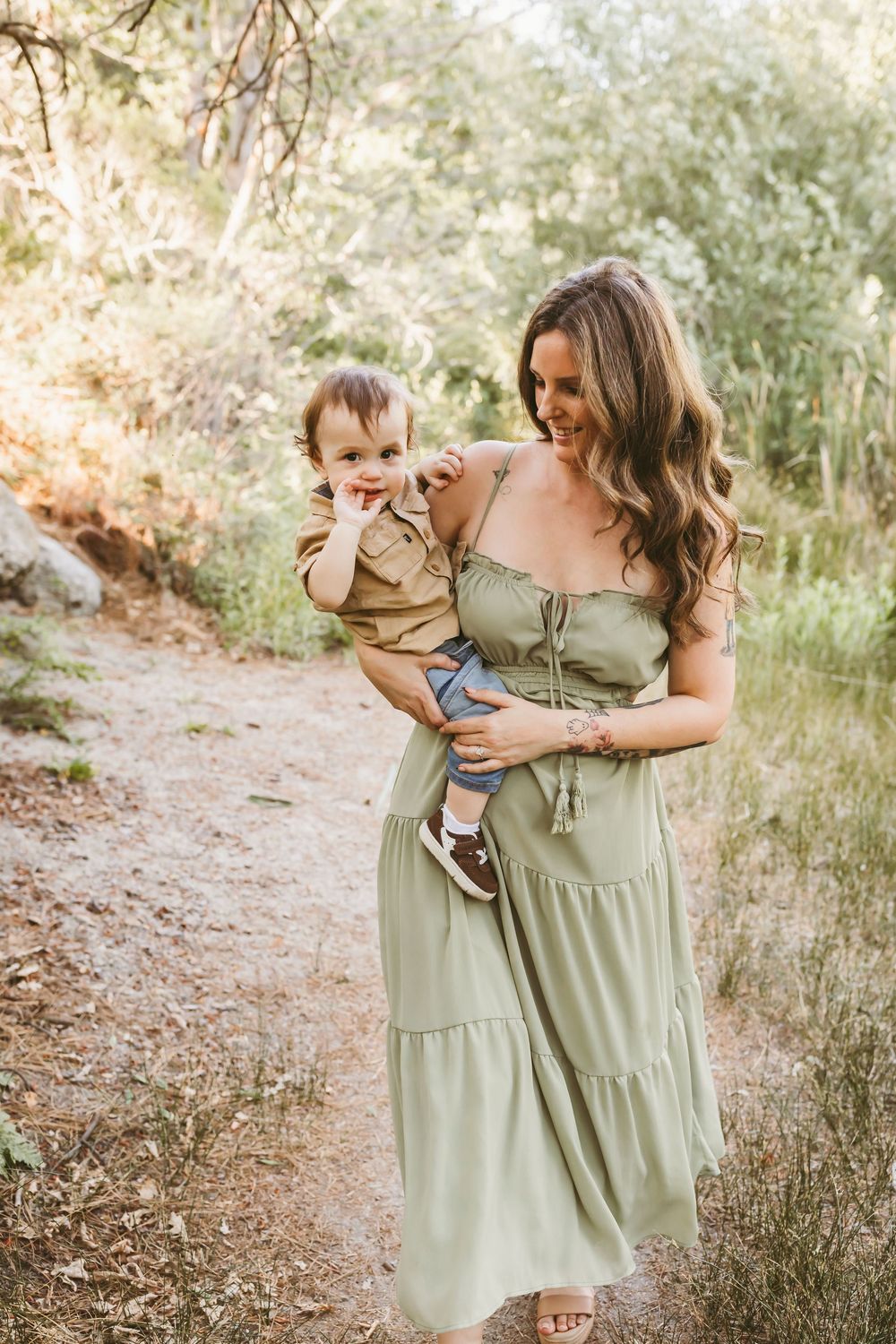 A smiling woman in a sage green dress holds a happy toddler on a sunlit forest path.