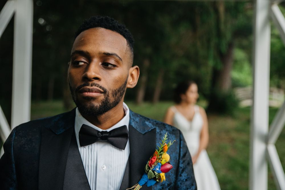 Groom in navy tuxedo and colorful boutonniere stands outdoors near garden foliage.