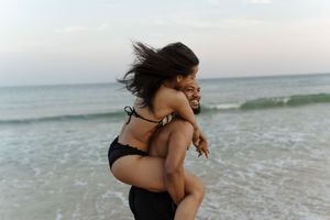 A joyful couple enjoys a piggyback ride on a sandy beach at dusk, with ocean waves in the background.