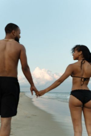 Couple holding hands on a sandy beach at dusk, man in black shorts and woman in black bikini.