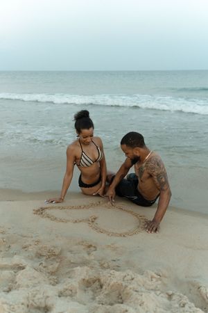 Couple sitting on sandy beach drawing a heart shape in the sand with ocean waves in the background.