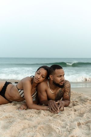 A couple relaxes on a sandy beach, the woman in a bikini leaning on a tattooed man wearing a gold chain.