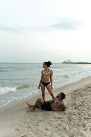 Couple on sandy beach at dusk, woman in striped bikini standing while tattooed man sits relaxing by ocean waves.