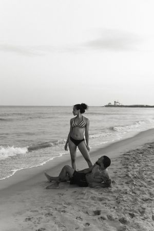 Black and white photo of a couple on a sandy beach, woman in bikini standing while man sits by the ocean waves.