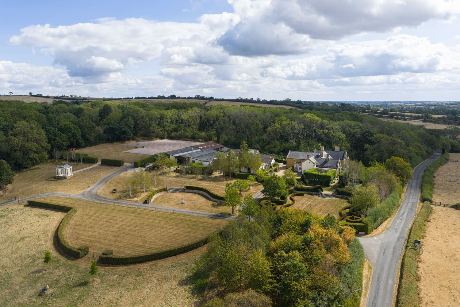 Aerial view of a rural estate with solar panels, manicured hedges, and rolling countryside alongside a quiet road.
