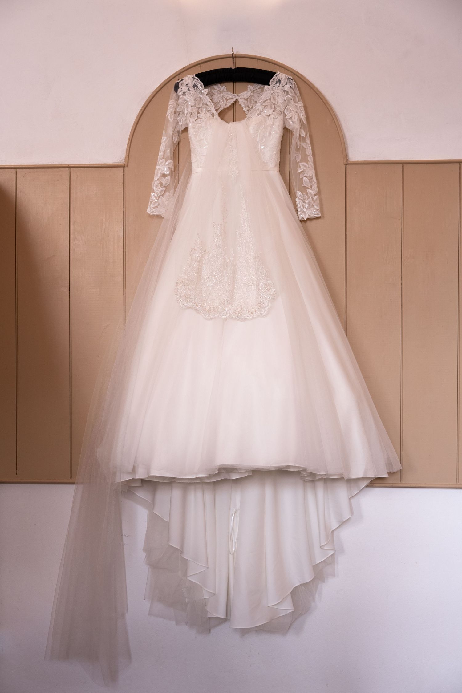 Elegant white lace wedding dress with long sleeves and flowing tulle skirt hanging on a wooden arch wall.