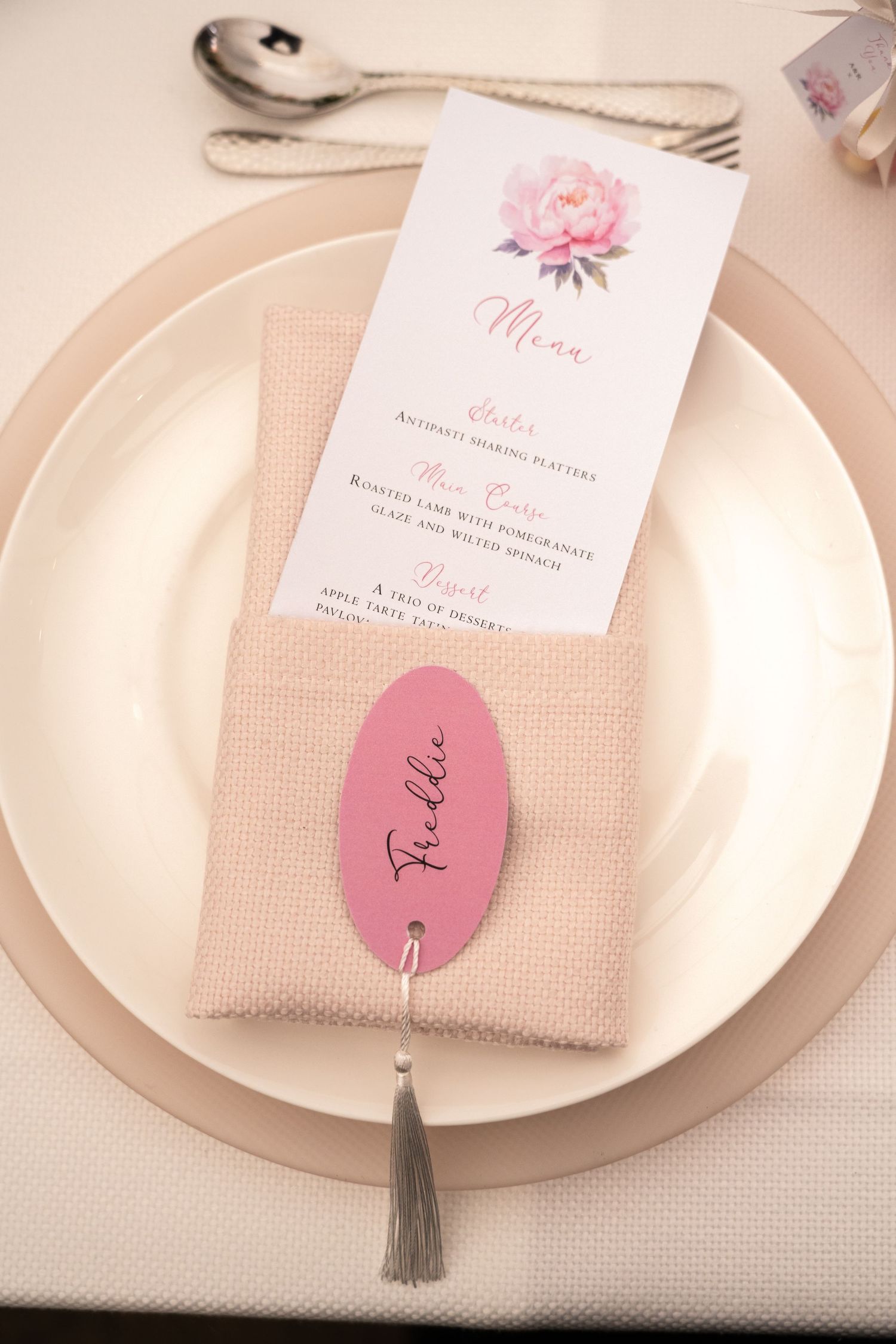 Elegant wedding place setting with pink peony menu card, burlap napkin holder, and oval pink name tag with tassel.