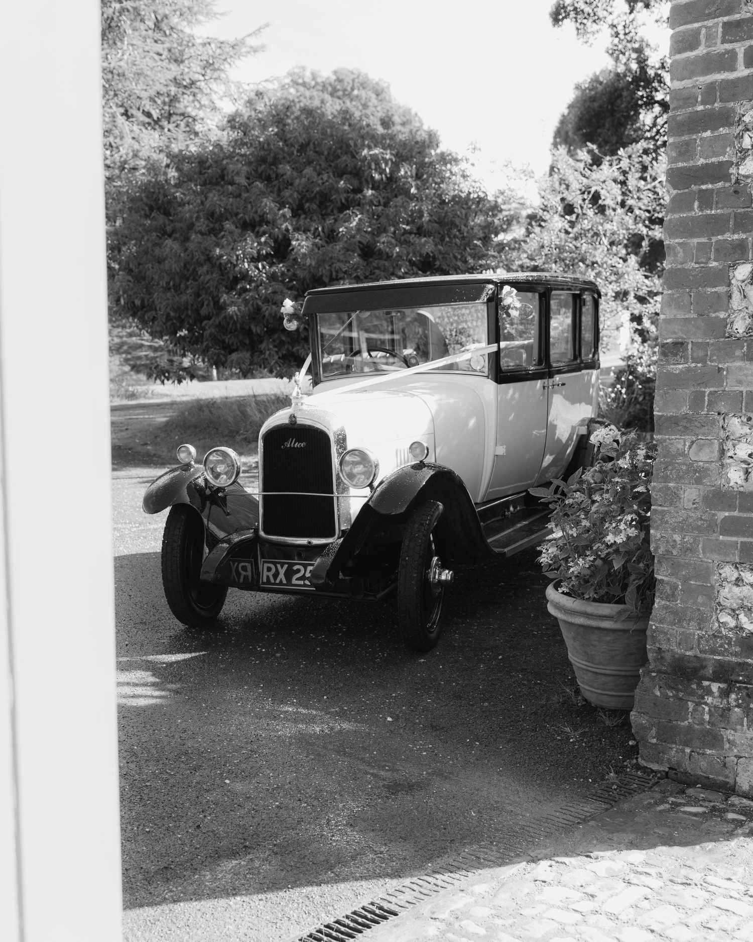 Vintage black and white photo of a classic antique car parked beside a brick wall, surrounded by lush trees.