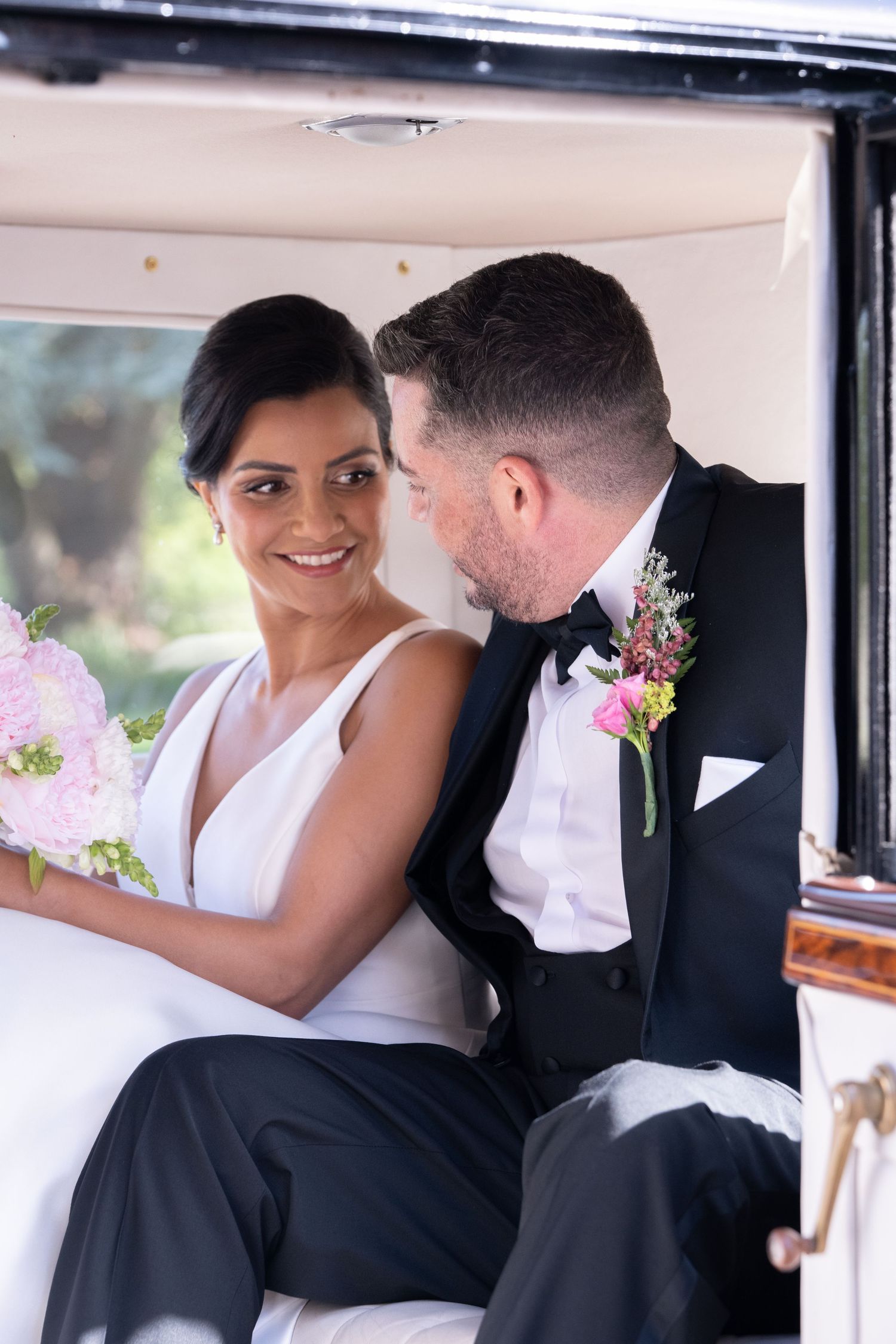 Bride and groom sitting in a vintage car, smiling at each other, bride holding pink bouquet, groom wearing boutonniere.