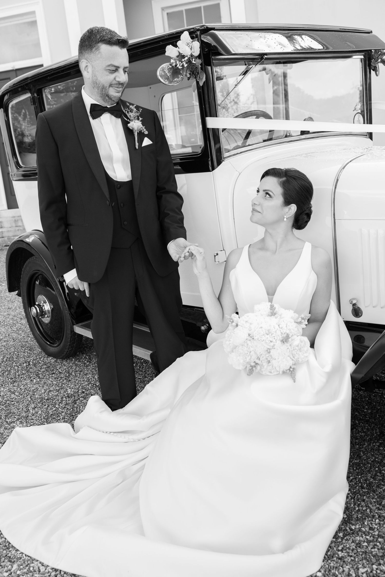 Black and white wedding photo of bride sitting in vintage car holding groom's hand, wearing elegant gown with bouquet.