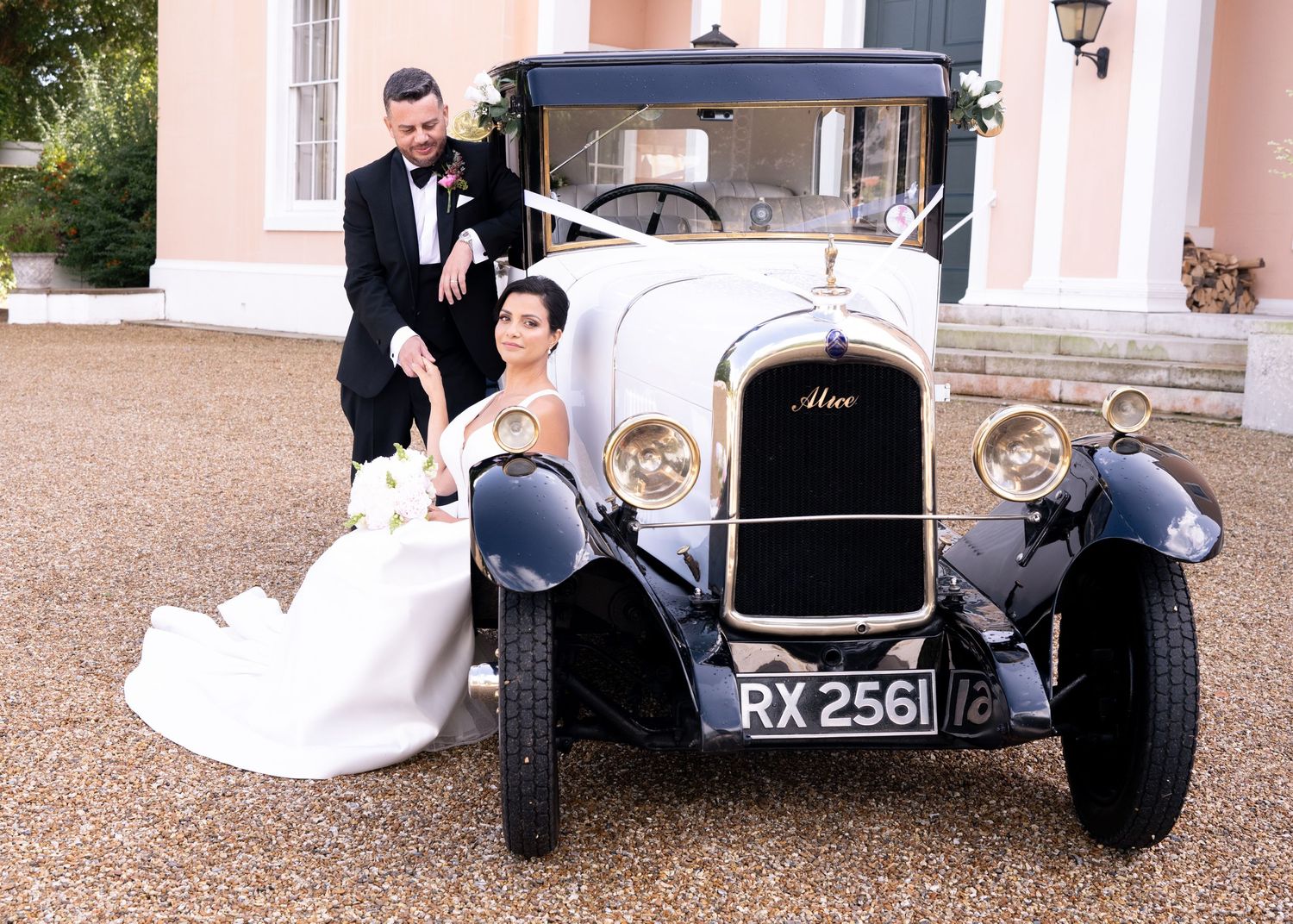 Bride and groom posing beside a vintage white and black classic car with RX 2561 plate at a wedding venue.