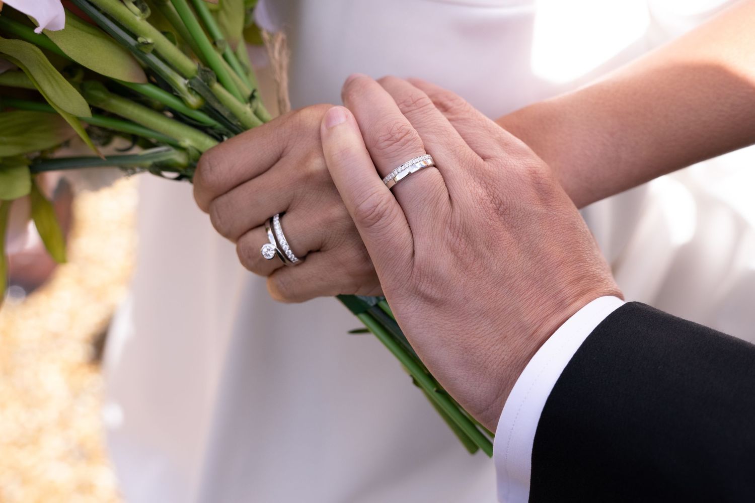 Newlywed couple holding a floral bouquet, showcasing matching diamond wedding bands on their ring fingers.