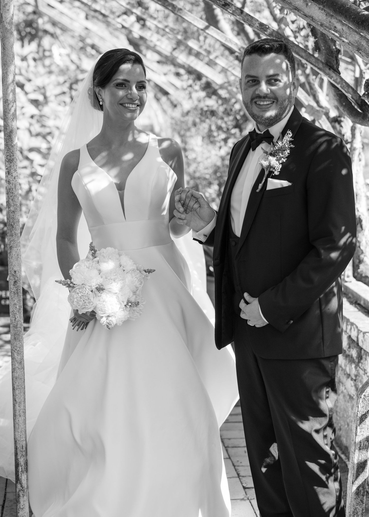 Black and white wedding photo of smiling bride in gown holding bouquet and groom in tuxedo standing together outdoors.