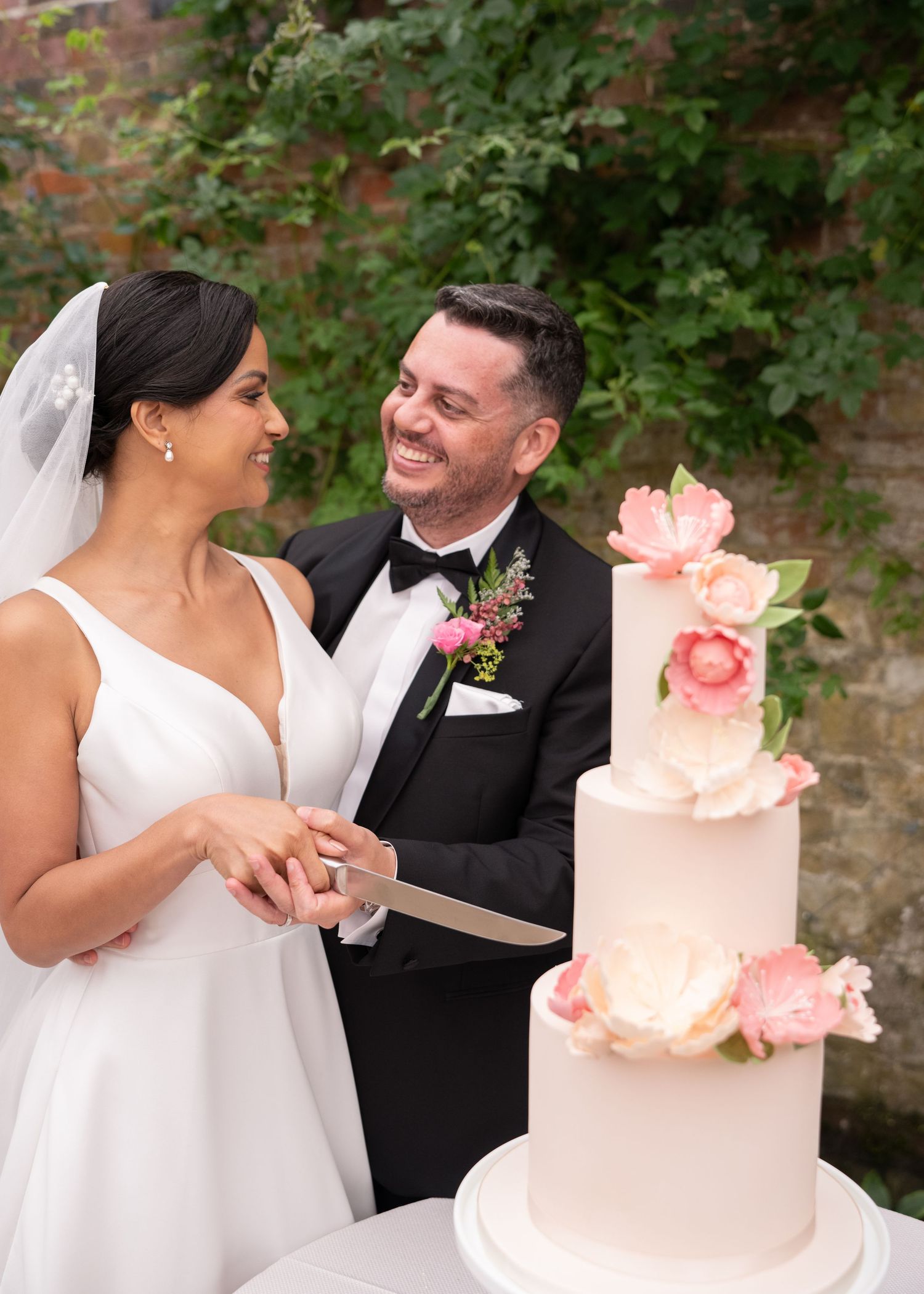 Bride and groom smiling while cutting a three-tier white wedding cake decorated with pink flowers outdoors.