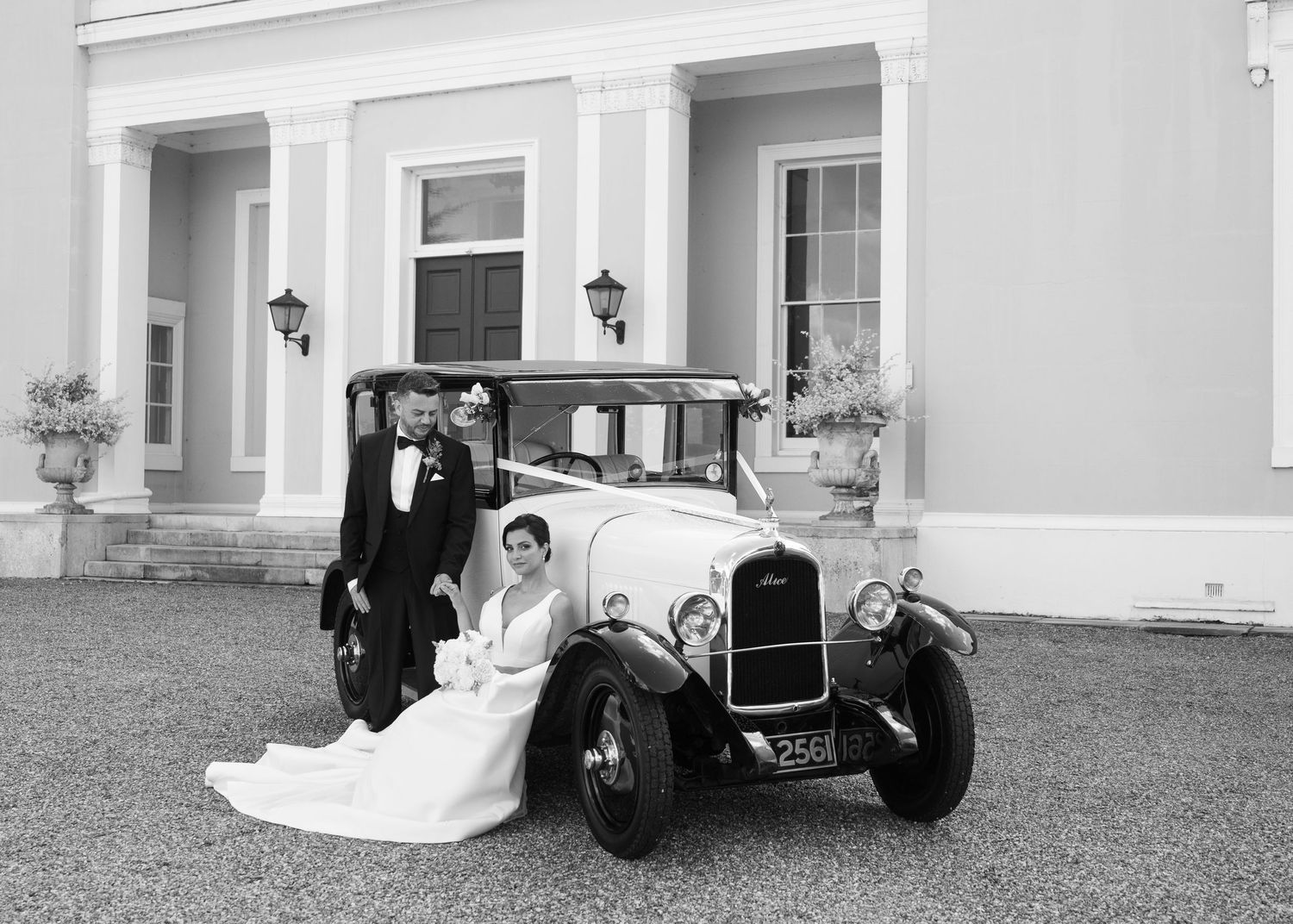 Black and white wedding photo of bride and groom posing with a vintage classic car outside an elegant manor house.