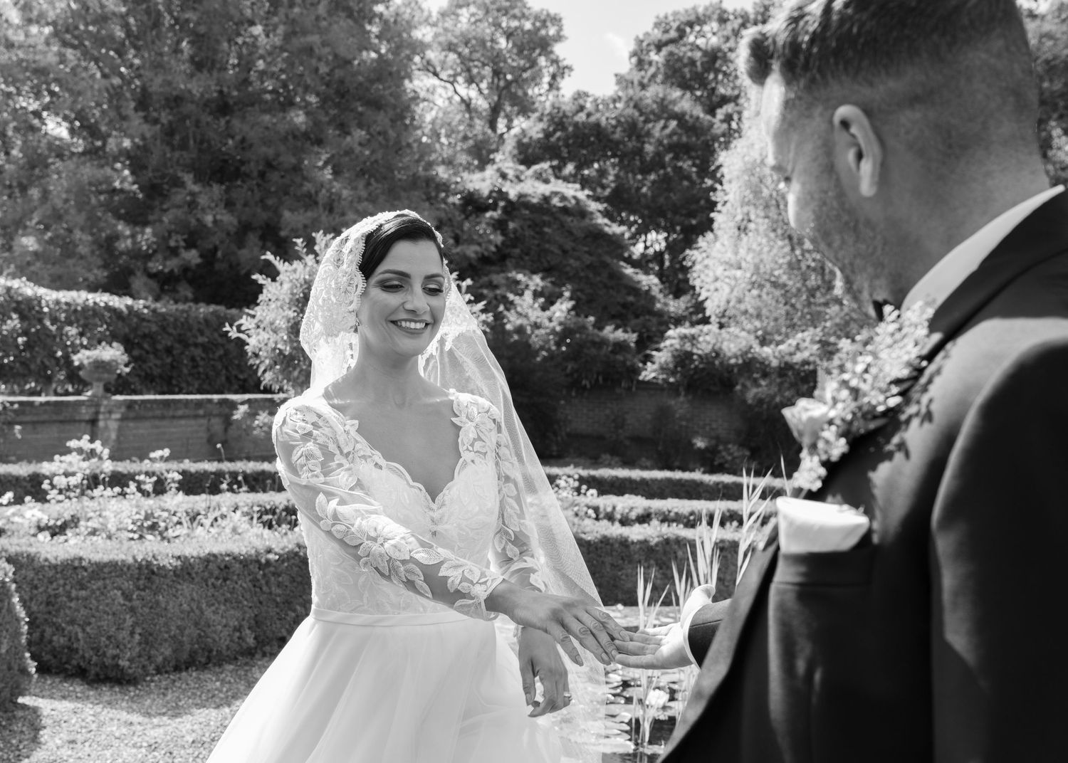 Black and white wedding photo of a smiling bride in lace gown and veil exchanging vows with groom in garden.