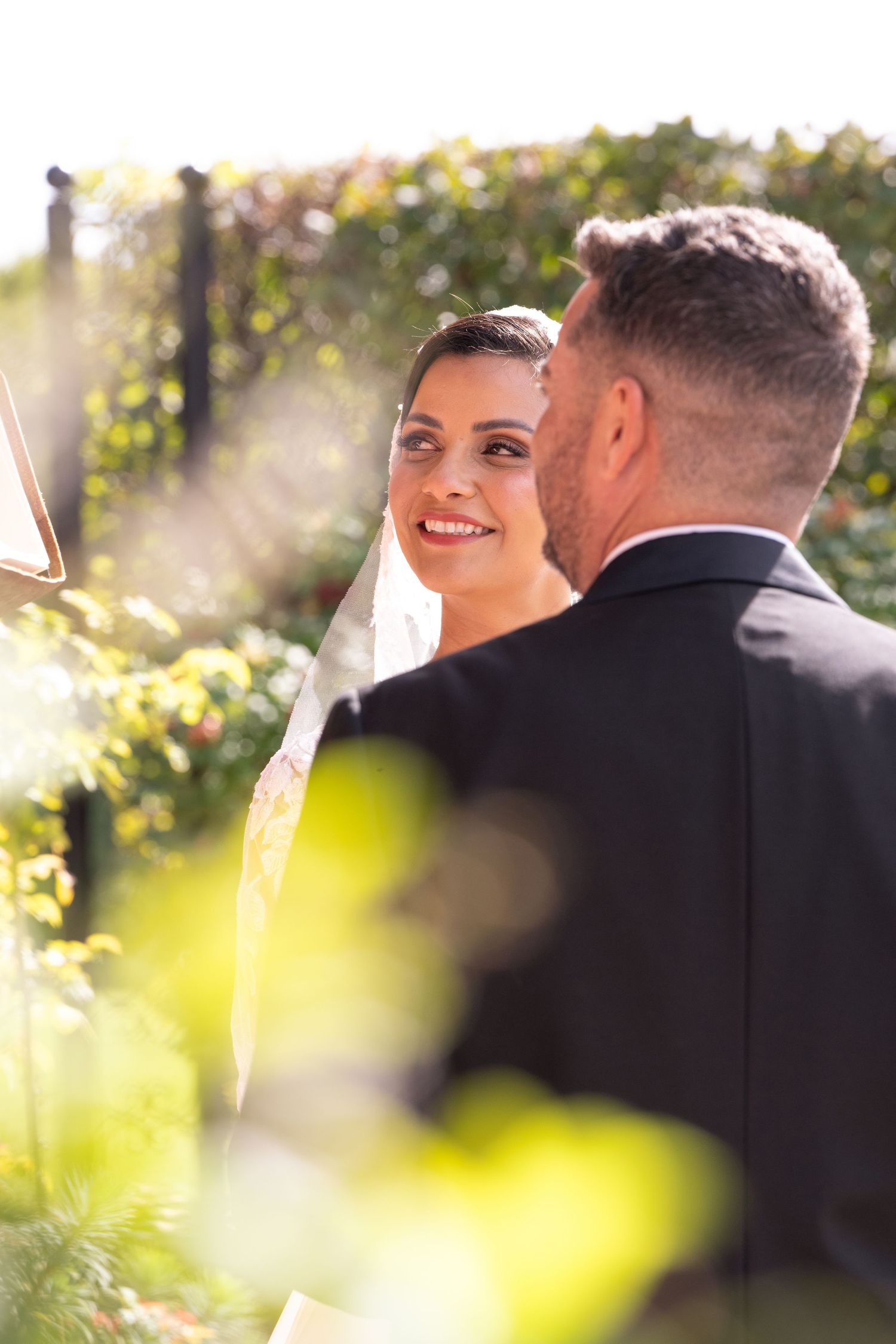Bride smiling at groom during outdoor wedding ceremony, bathed in warm sunlight with lush greenery backdrop.