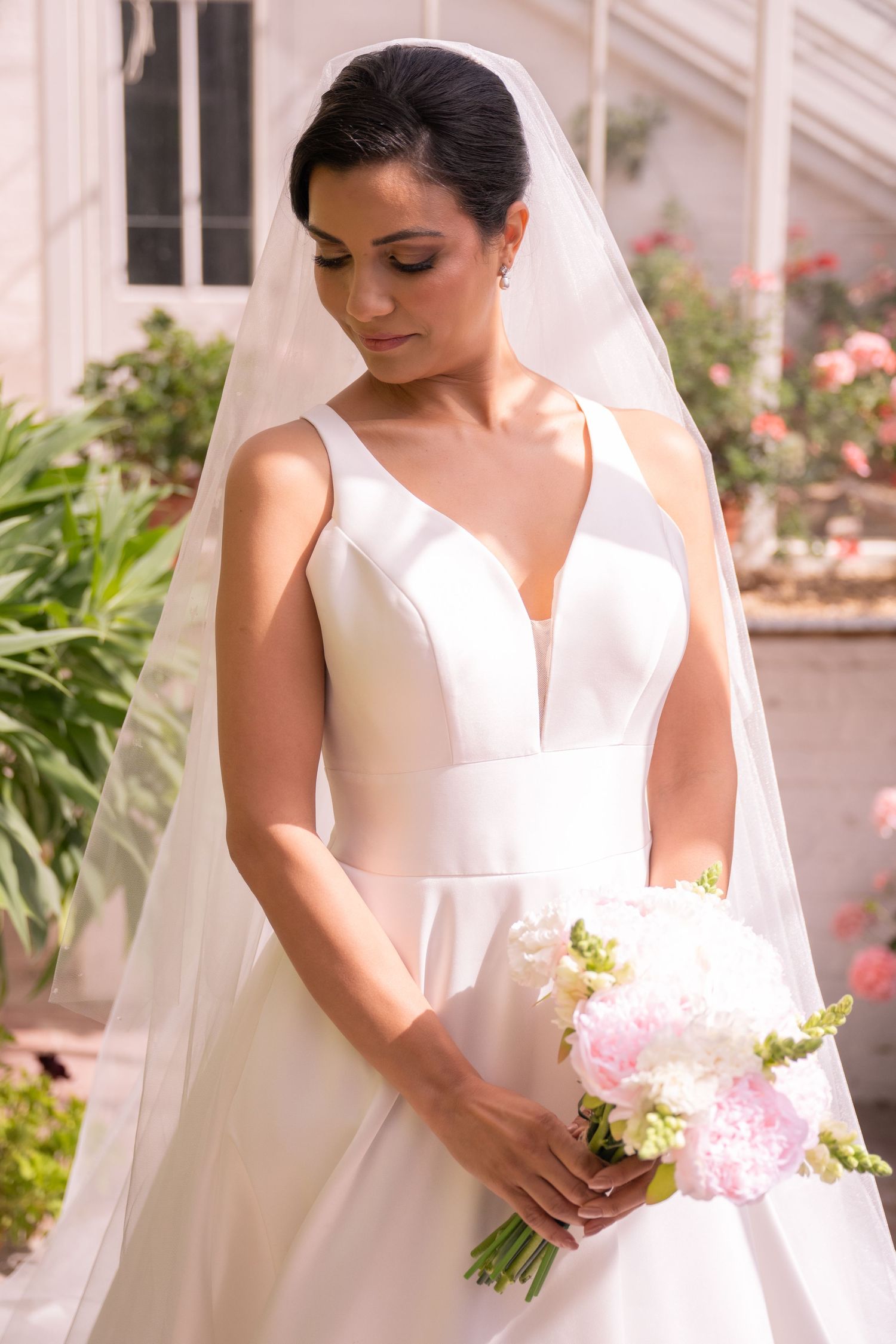 Bride in white V-neck ball gown and veil holds pink and white bouquet, standing outdoors near roses.