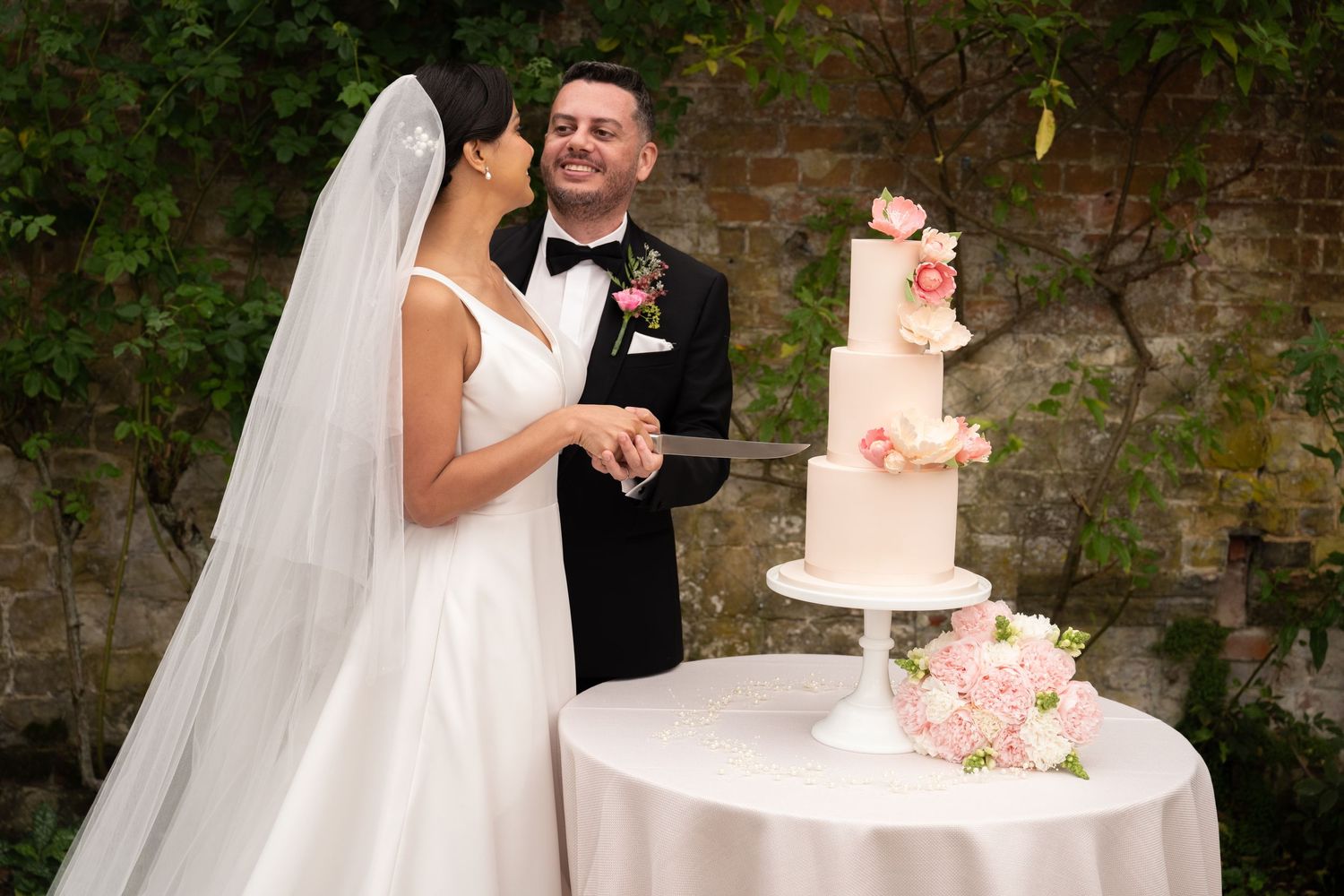 Bride and groom cutting a three-tier white wedding cake with pink roses outdoors at their elegant wedding reception.