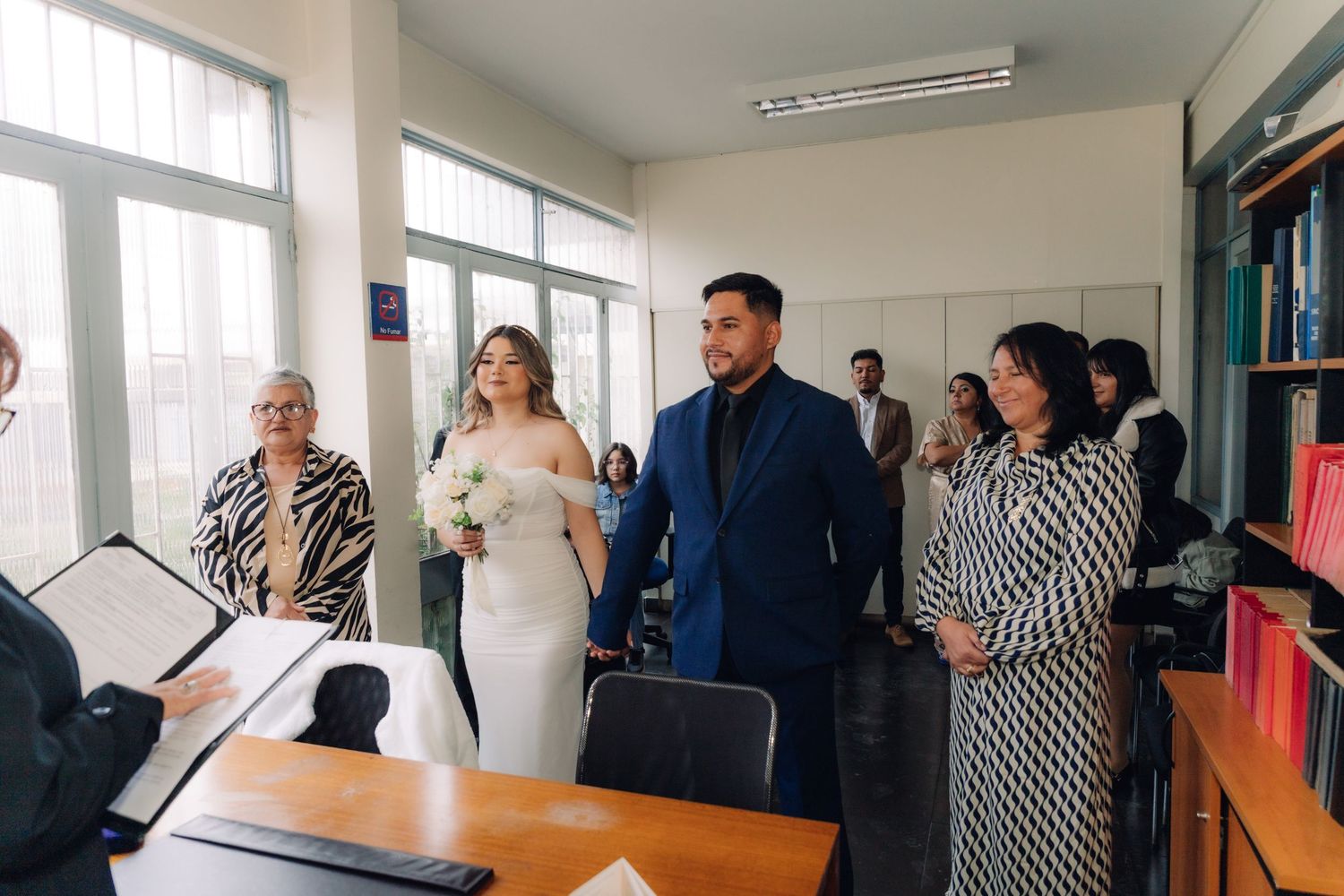 Wedding couple holding hands at a civil ceremony office, surrounded by guests and an officiant.