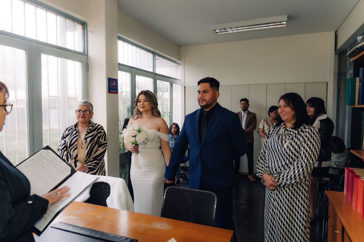 Bride in white off-shoulder gown and groom in navy suit stand before officiant during civil wedding ceremony.