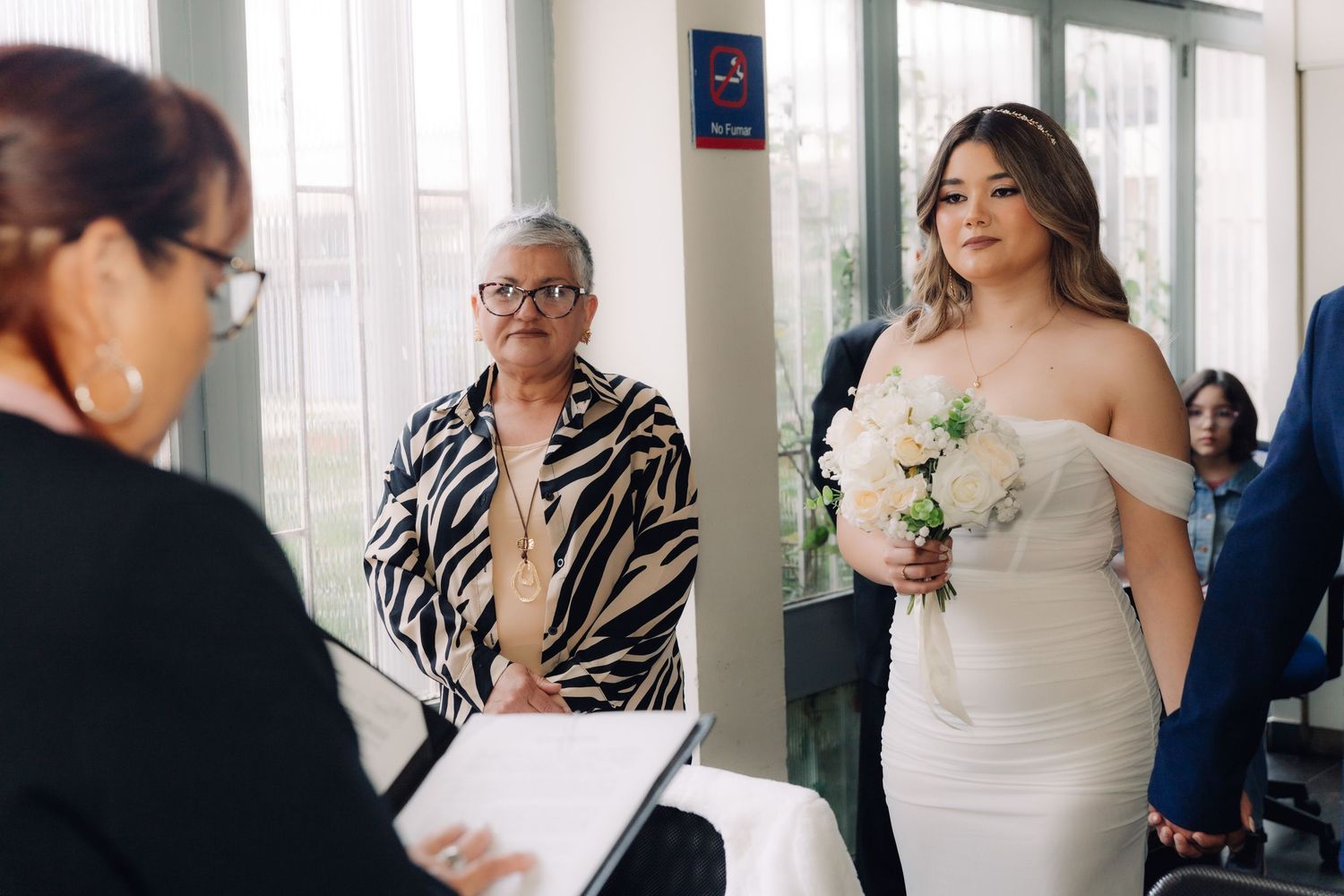 Bride holding white rose bouquet stands beside older woman in zebra print jacket during wedding ceremony.