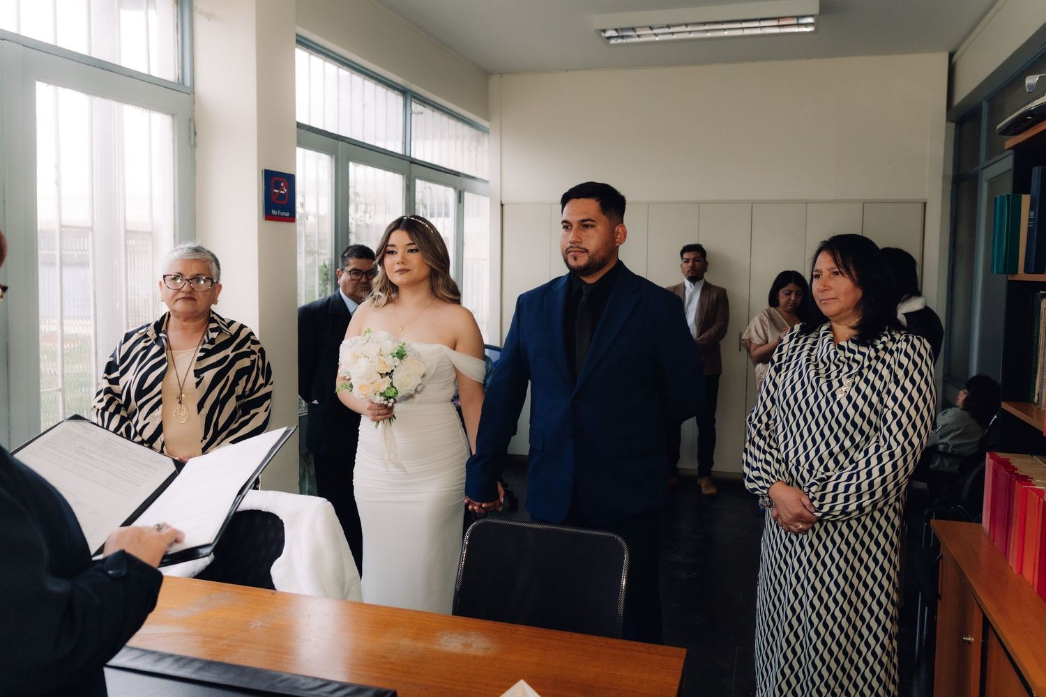 Couple holding hands before officiant with wedding guests standing alongside during indoor civil ceremony.