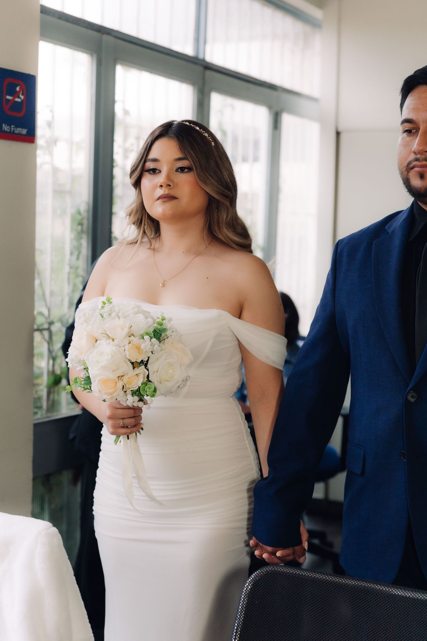 Bride in white off-shoulder fitted gown holds white rose bouquet while holding grooms hand at ceremony.