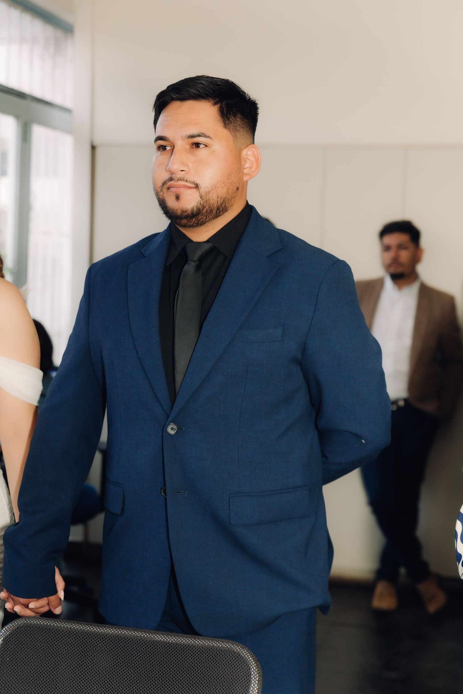 Groom in navy blue suit with black shirt stands attentively during wedding ceremony indoors.