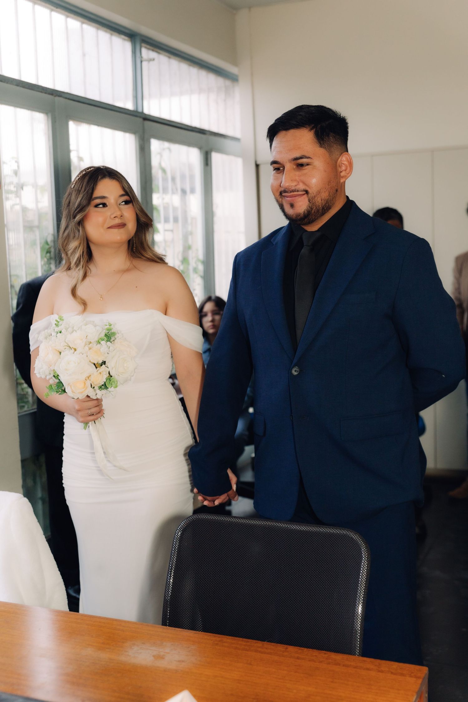 Smiling bride and groom hold hands standing before a desk during their civil wedding ceremony.