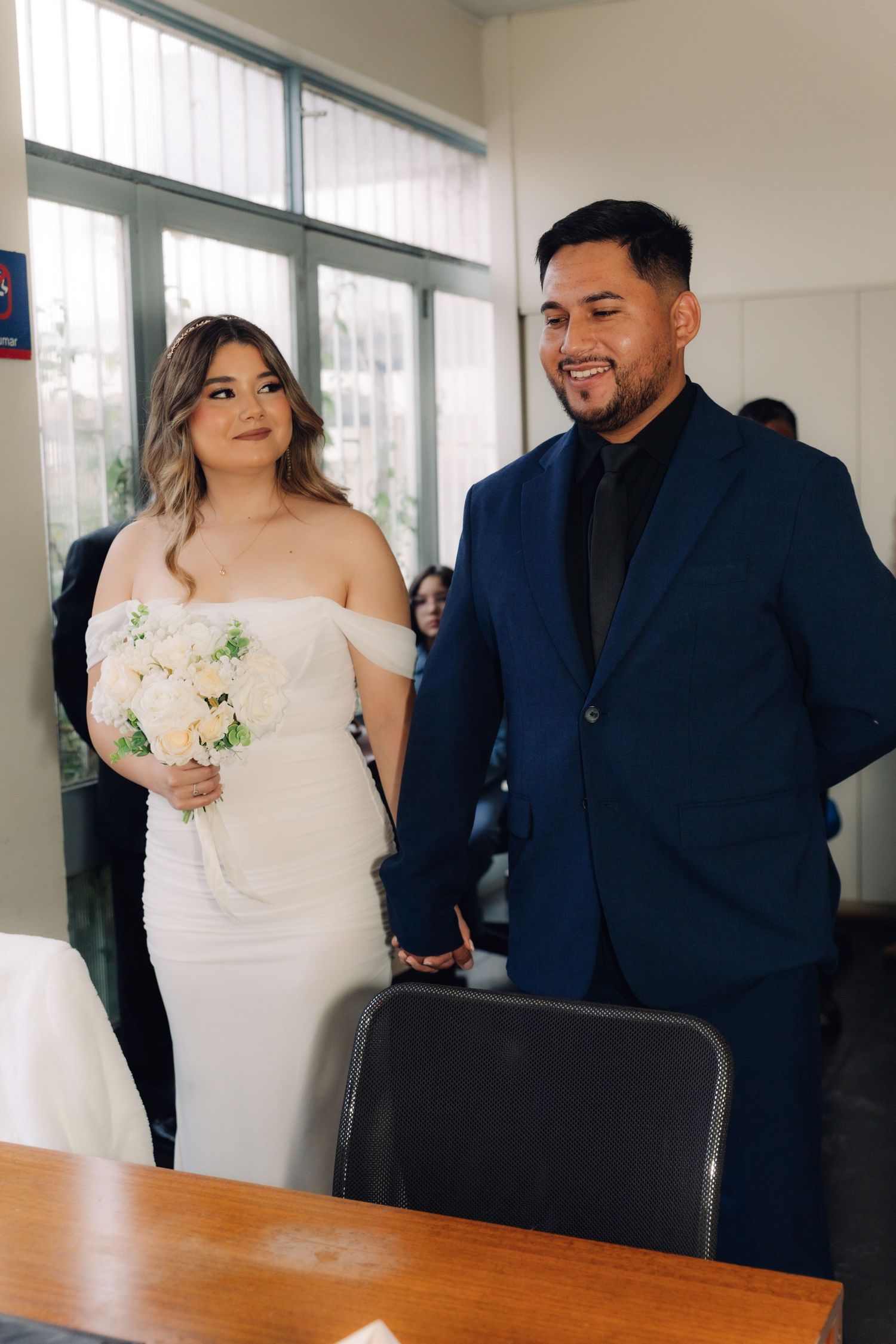 Bride with white bouquet and groom in navy suit smile while holding hands at civil ceremony.