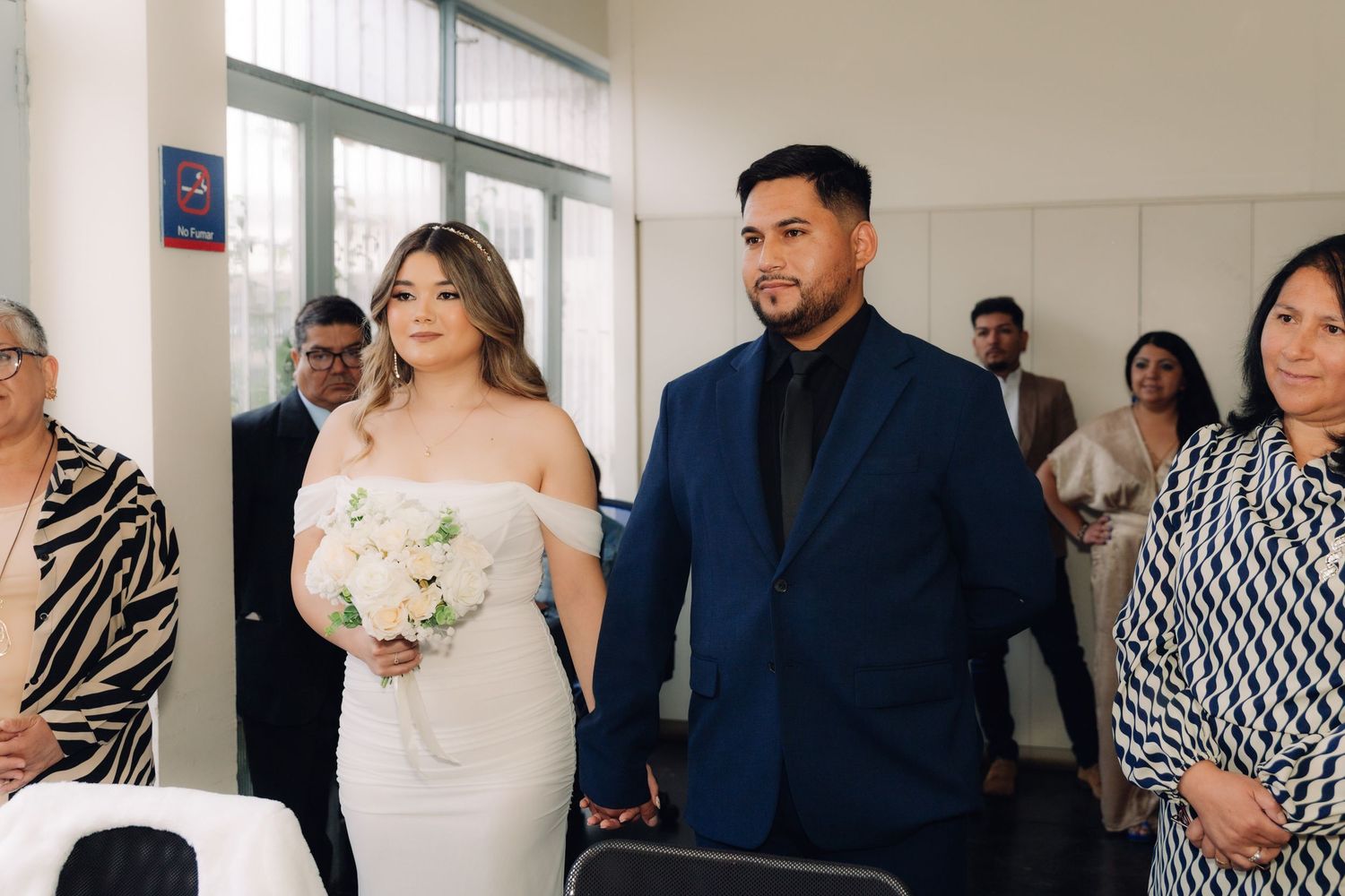 Bride holding white rose bouquet and groom in navy suit hold hands during civil wedding ceremony.